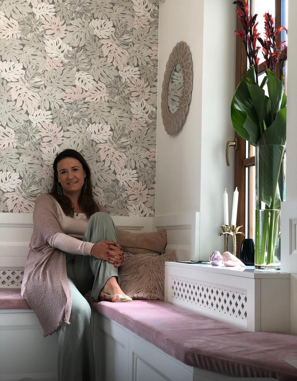 Dr. Kamila Hortyńska sitting on a pink velvet bench in a stylish interior. She is leaning against a wall with tropical leaf wallpaper, surrounded by soft cushions and greenery.